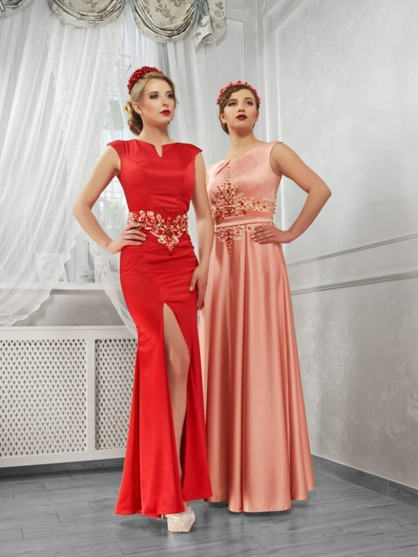 Two young, beautiful women in long, evening red and peach dresses standing shoulder to shoulder on hills on white background in the room.